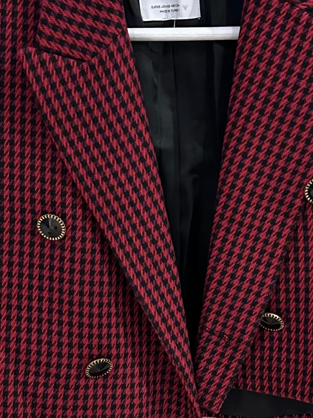 Close-up of a red and black houndstooth jacket with buttons. Text: EUR XS USA XS MEX 24 MADE IN TURKEY.
