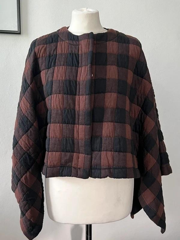 Short padded jacket in brown and black check pattern with wide sleeves, displayed on a white mannequin in front of a light-coloured wall.