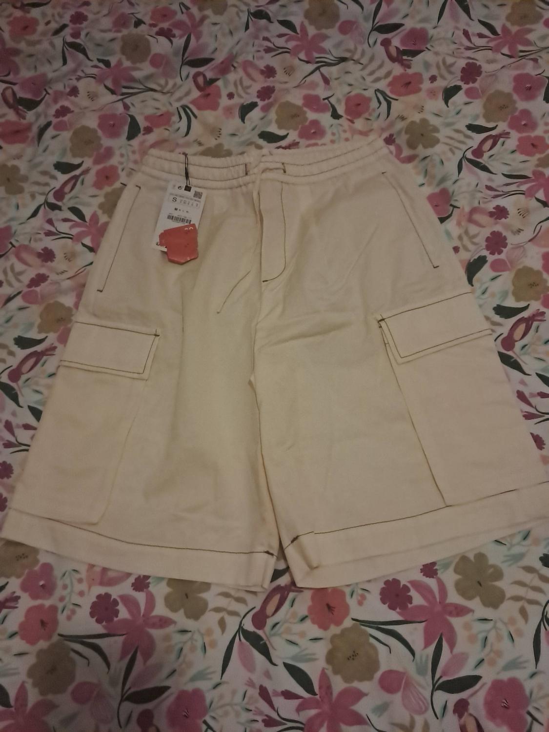 Beige cargo shorts with an elasticated waist, drawstring and side pockets, against a fabric background with a floral print.