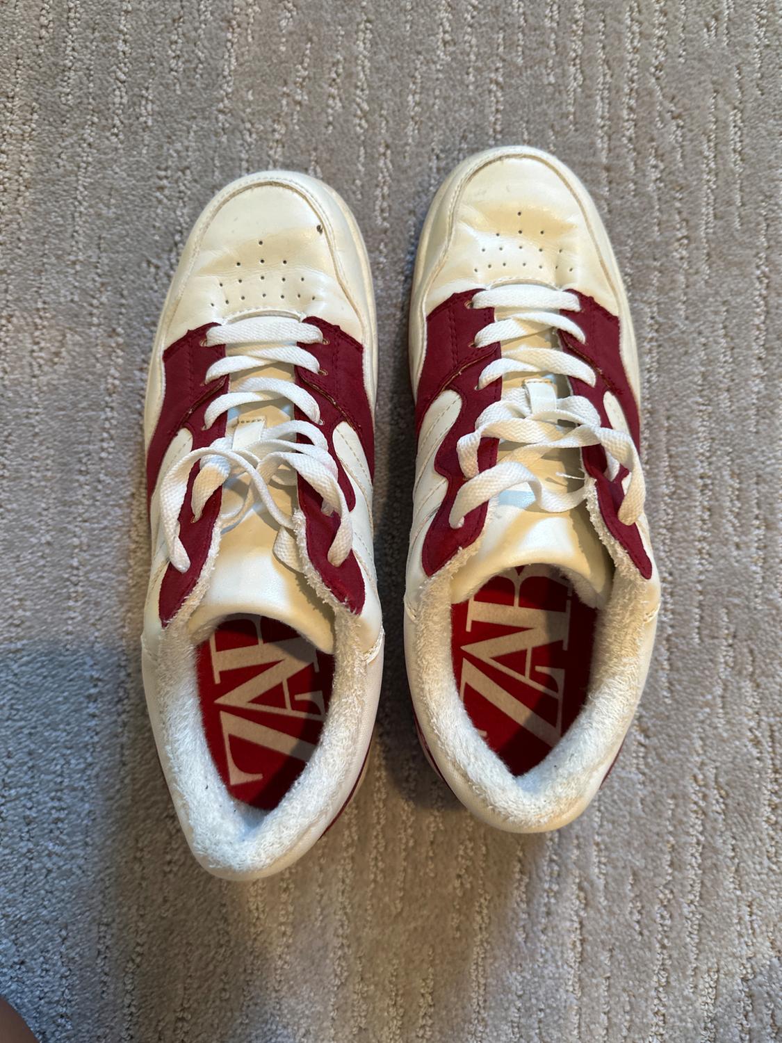 White athletic shoes with red details and white laces on a gray carpet. Insoles with the text "ZAP".
