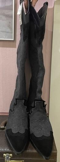 Black knee-high boots with decorative details, hanging on a pink wall. Background with a photo frame and a brown suitcase.