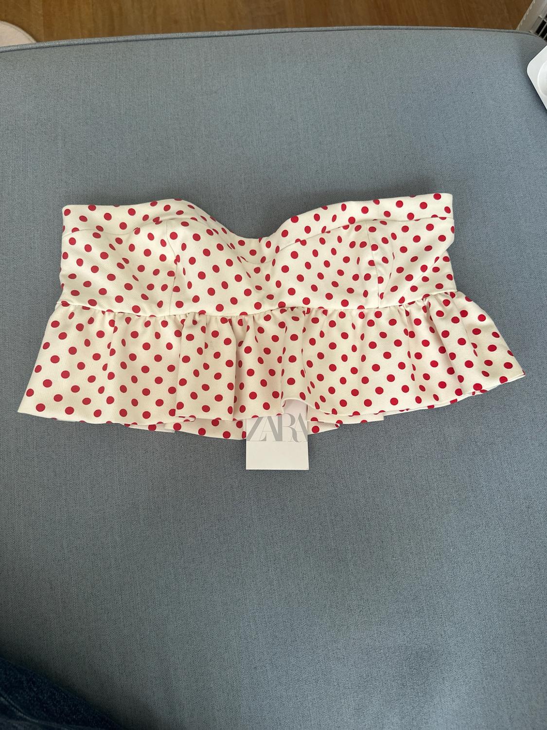 White strapless top with red polka dots and a ruffled hem, on a blue fabric surface. Label with the text ZARA.