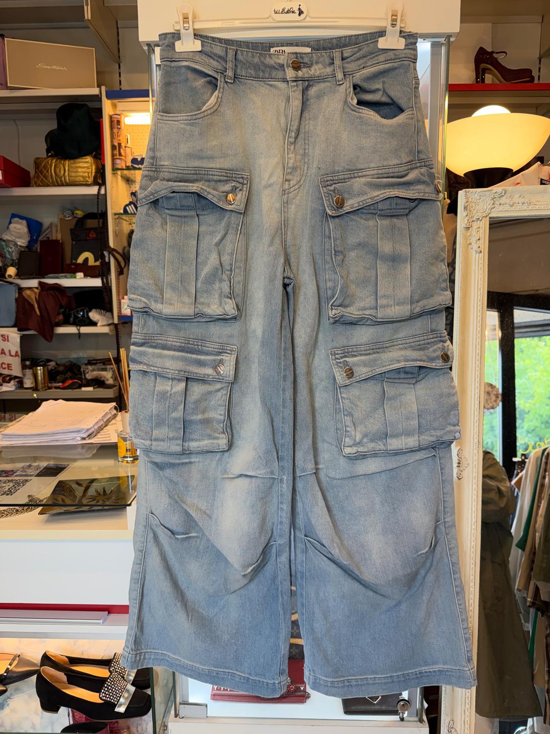Light blue denim cargo trousers hanging on a hanger, with large pockets and buttons, in a shop with shelves and shoes.