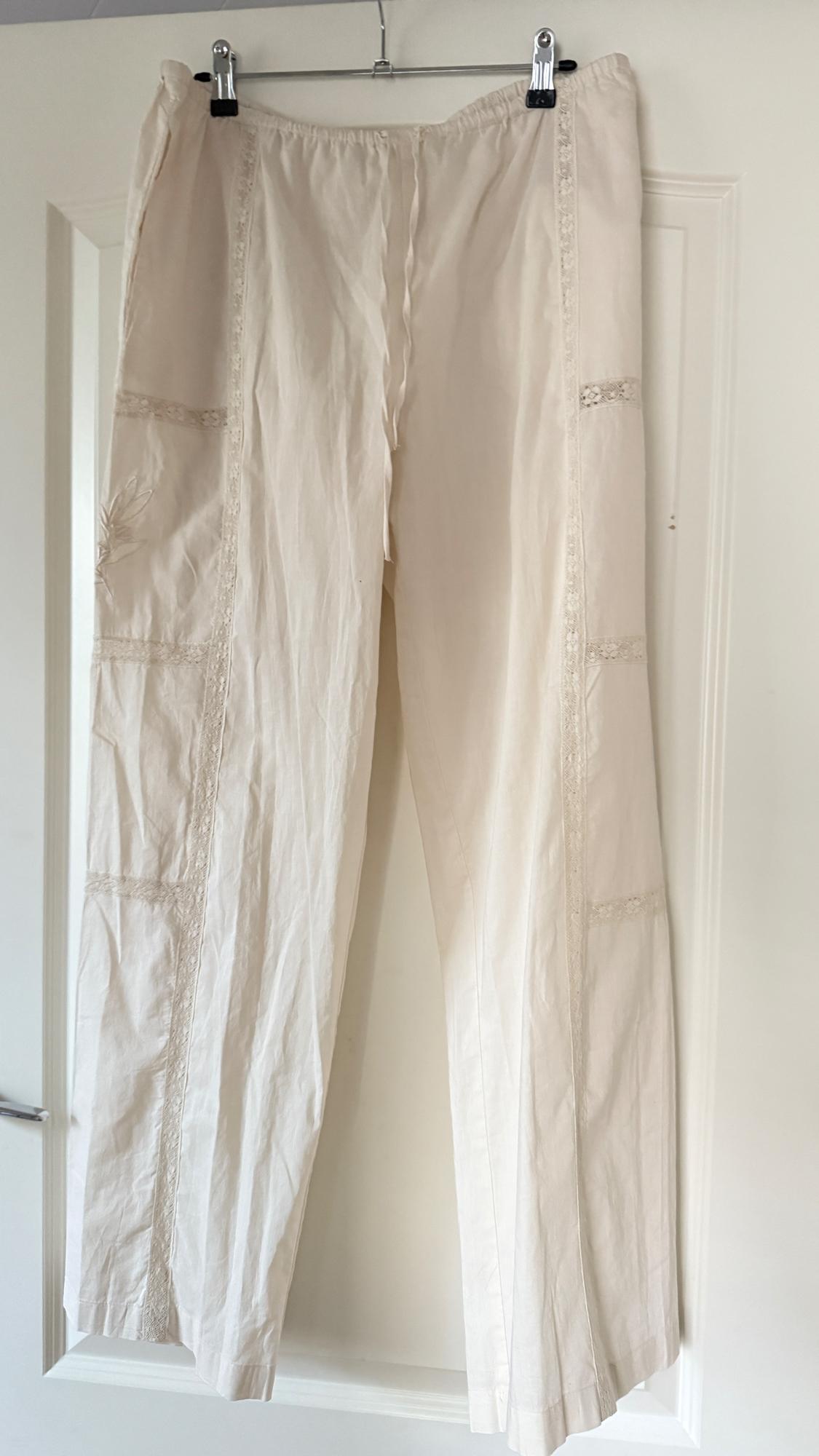 Beige trousers hanging on a hanger, with lace detailing on the seams and floral embroidery on the left leg. White door background.