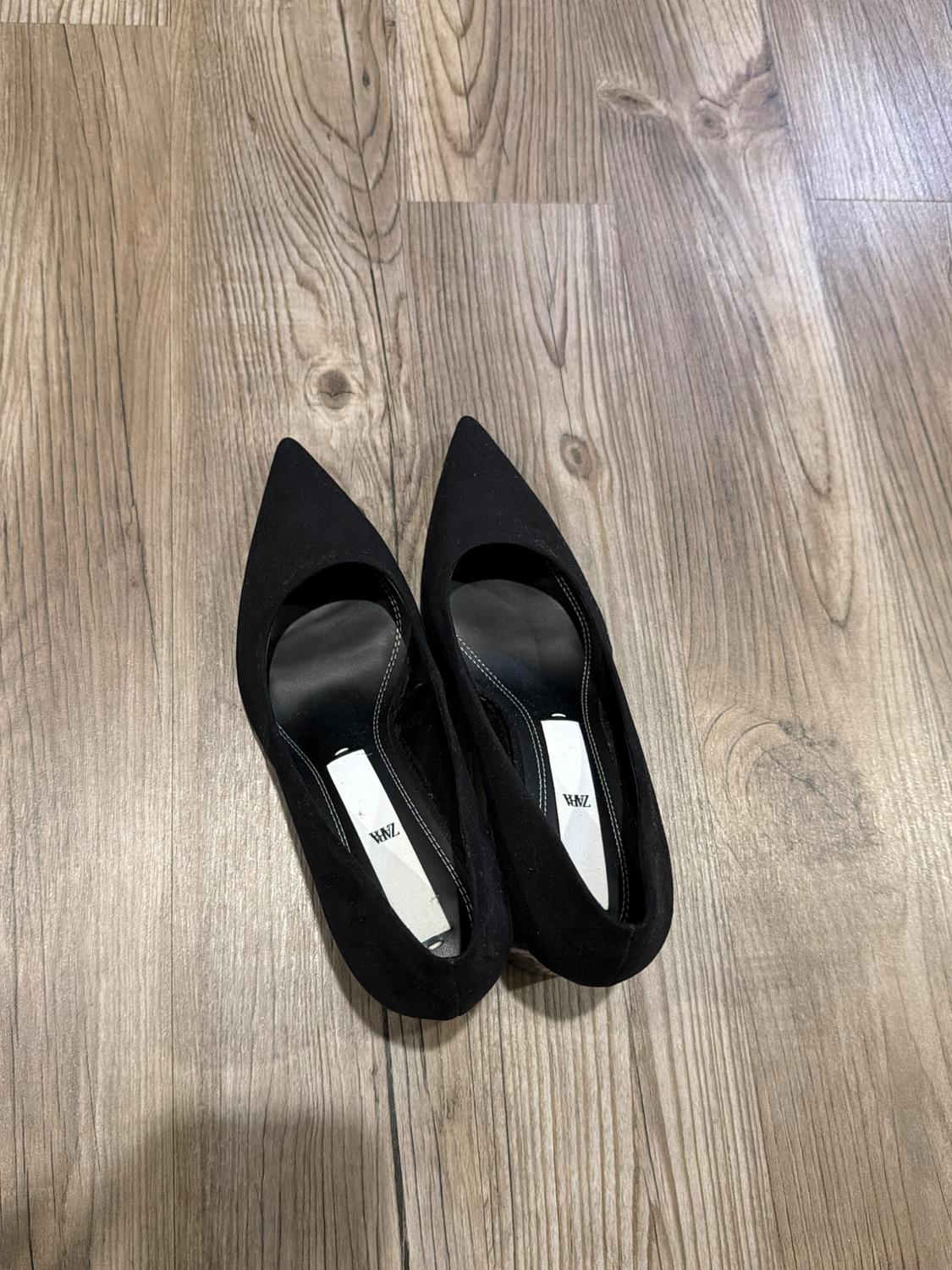 Black pointed-toe high heels on a wooden floor. “ZARA” is visible on the white insole.