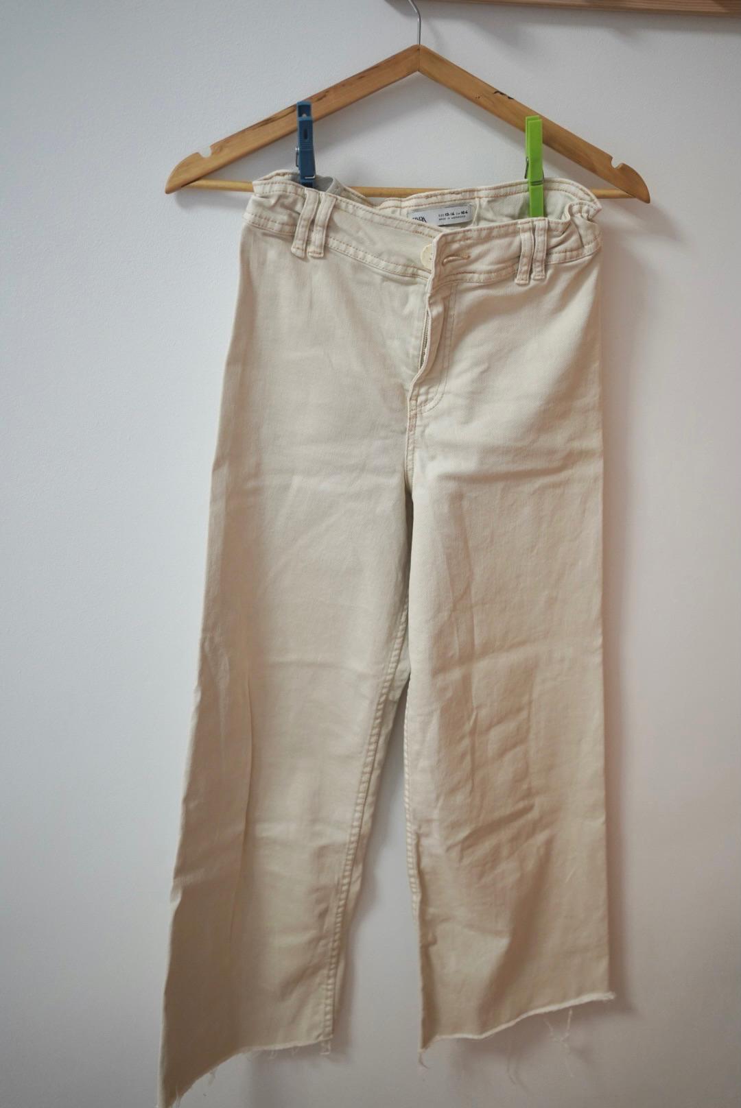 Beige trousers hanging on a wooden hanger with blue and green pegs. Plain white background.
