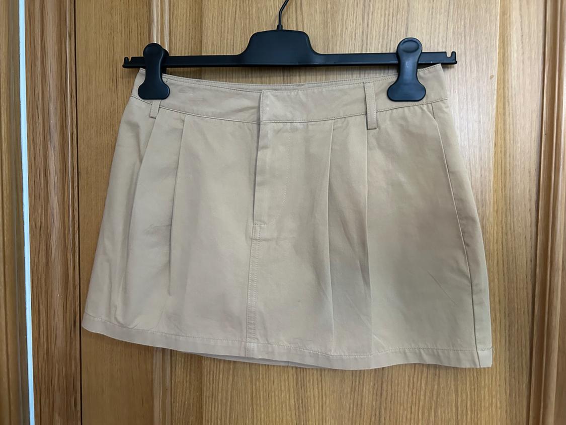 Beige mini skirt with darts and belt loops, hanging on a black hanger over a light wooden door. Casual style.