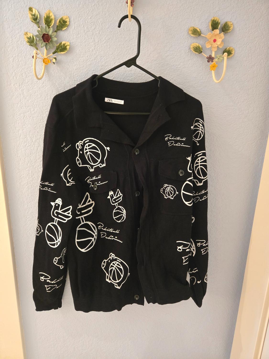 Black shirt with white drawings of pigs and footballs, hanging on a black hanger against a light blue wall with decorative hooks.