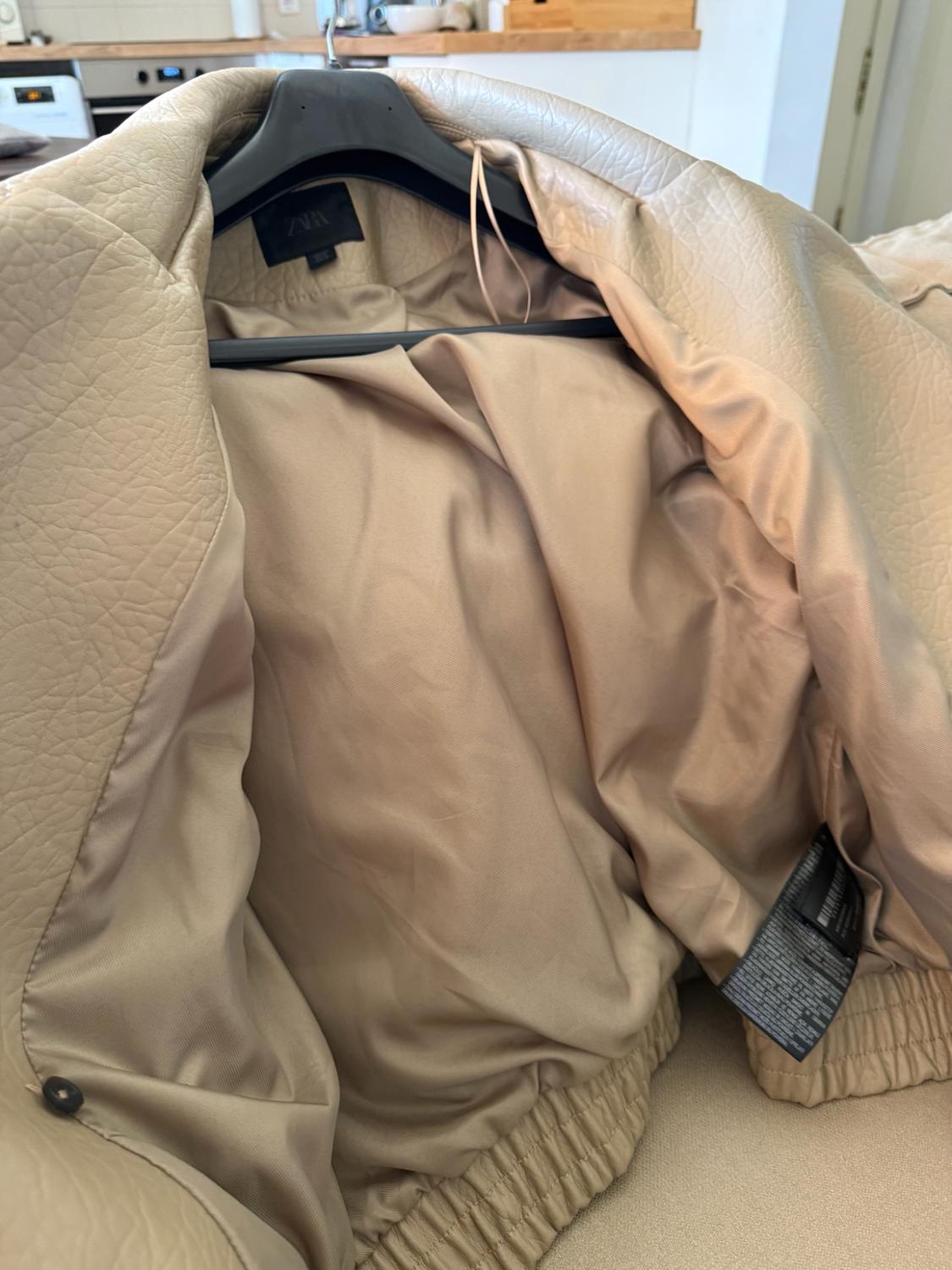 Beige jacket hanging on a black hanger, open to show its inner lining and the ZARA label. Kitchen background.
