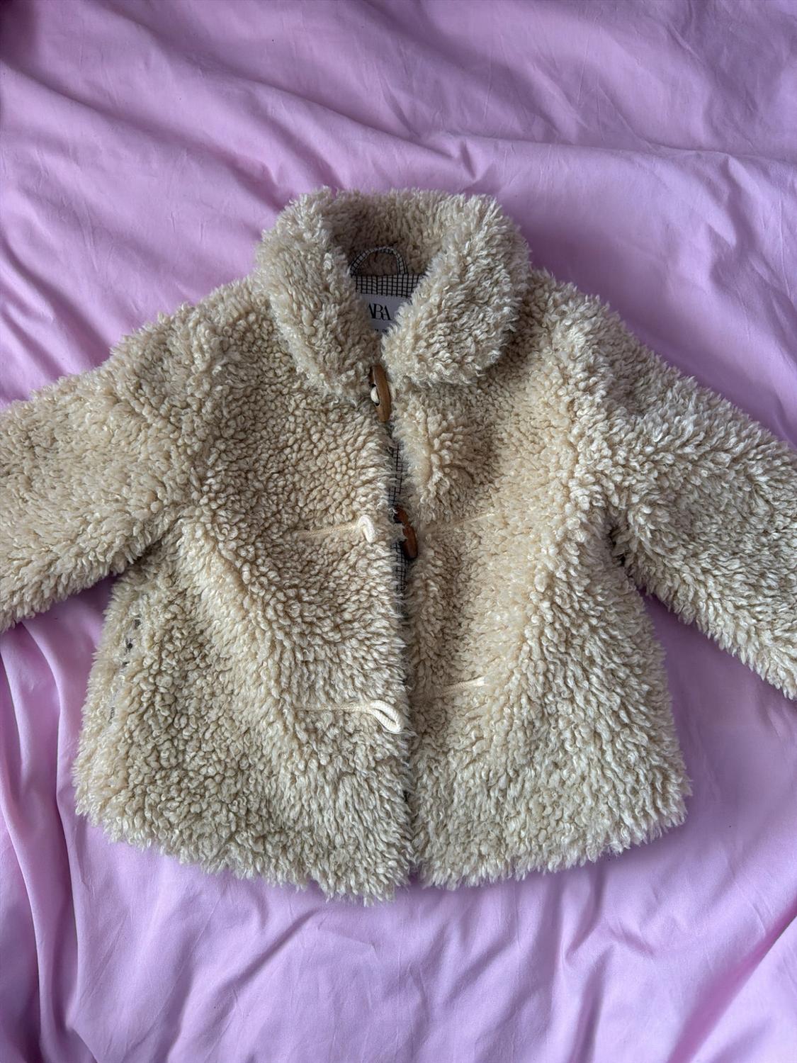 Beige curly faux-fur jacket with wooden buttons, resting on a crumpled pink sheet. Inner label featuring the text Zara.