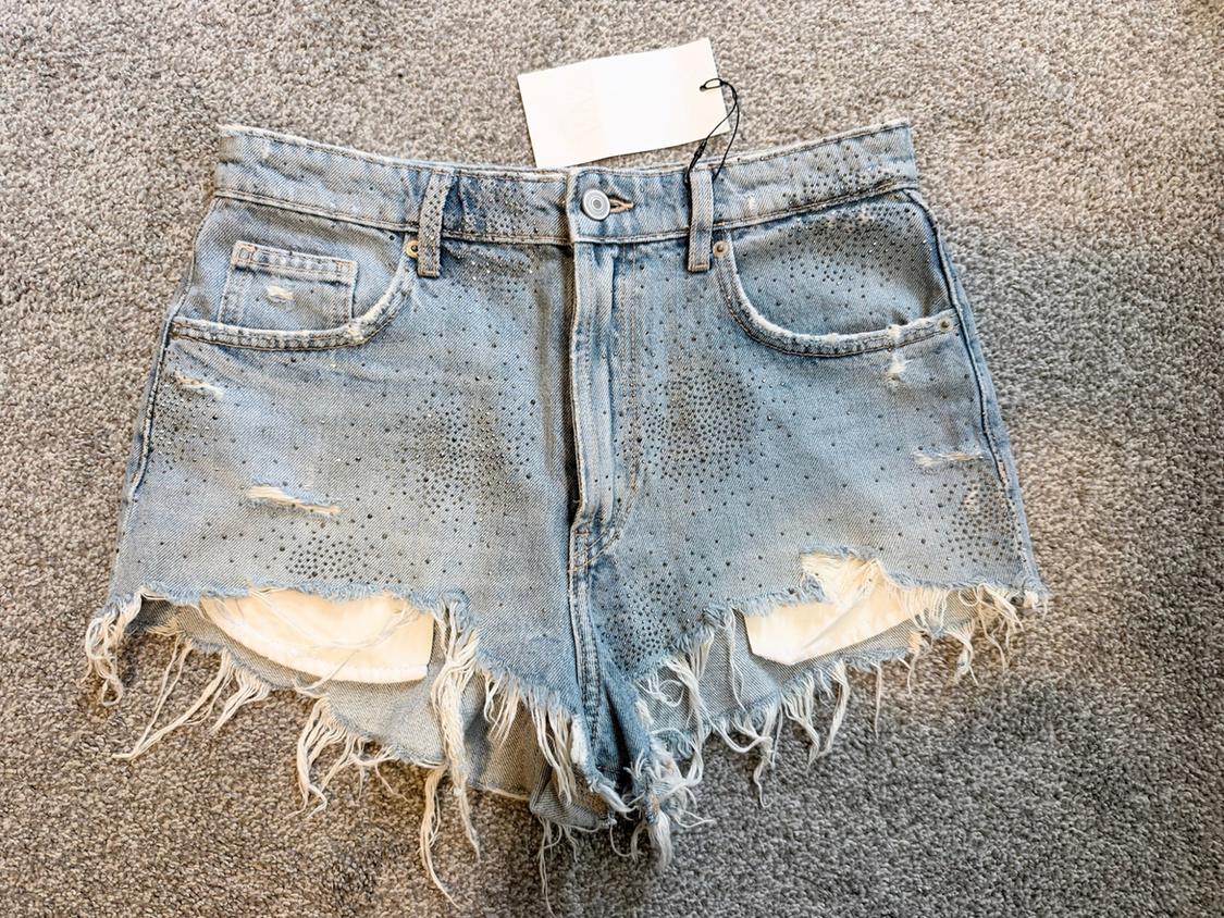Light blue denim shorts with rhinestone embellishment, distressed rips, and frayed hems that show the pockets, on a gray rug.