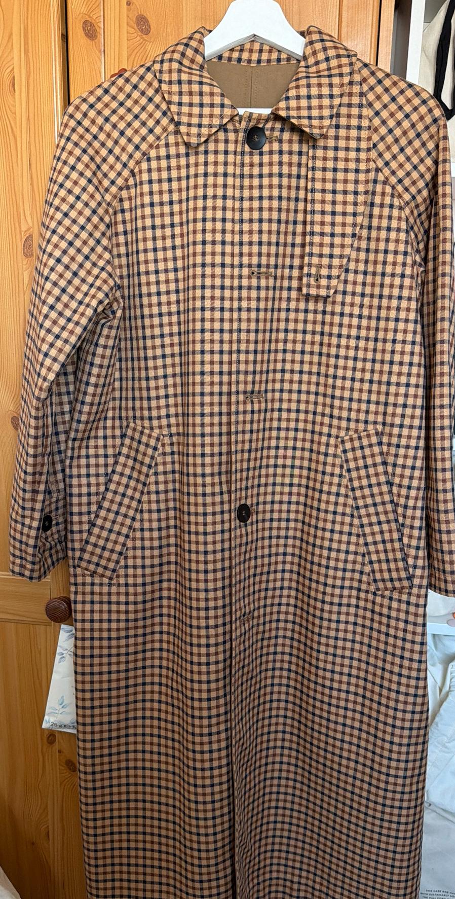 Long check-pattern coat in earthy tones and black, with dark buttons and pockets, hung on a white hanger in front of a wooden wardrobe.