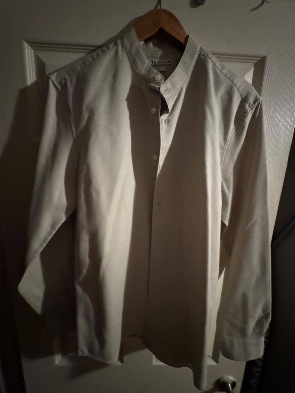 Beige long-sleeved shirt hanging on a wooden hanger over a white door, with dim lighting.
