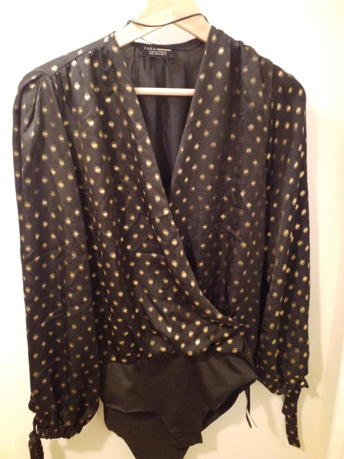 Black blouse with gold polka dot print, long sleeves and a wrap-over neckline, hanging on a wooden hanger.