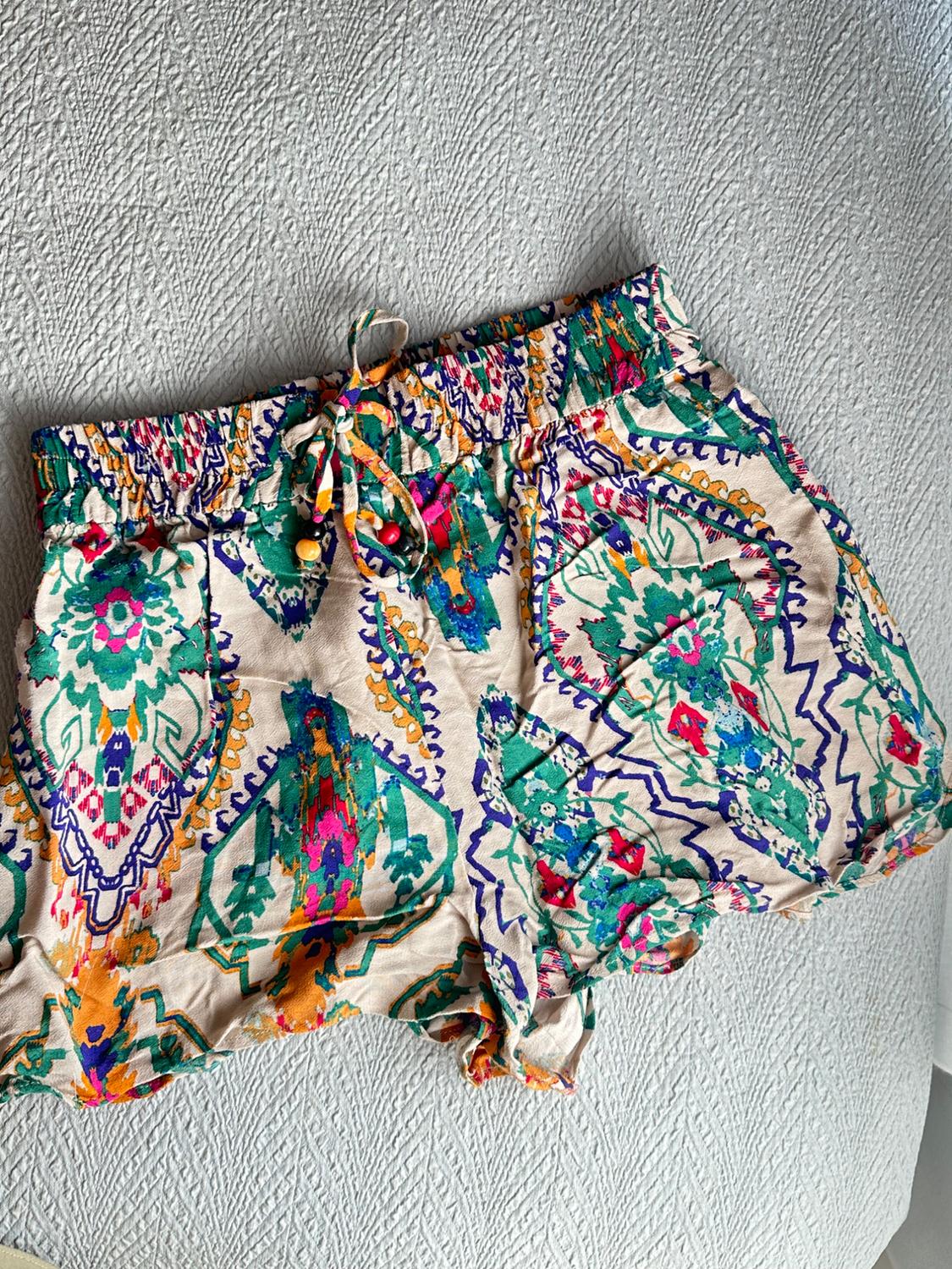 Lightweight beige shorts with a multicoloured geometric print. Elasticated waist with a drawstring and beads, on a textured grey background.