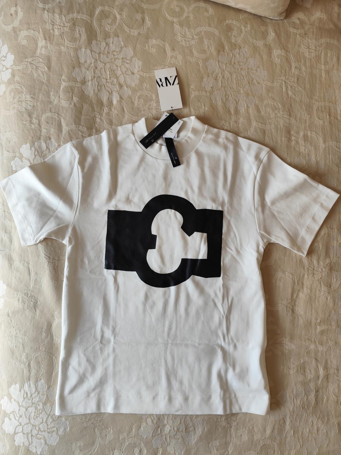 White T-shirt with an abstract black design in the centre, on a beige floral bedspread. Visible label with "ZARA" text.