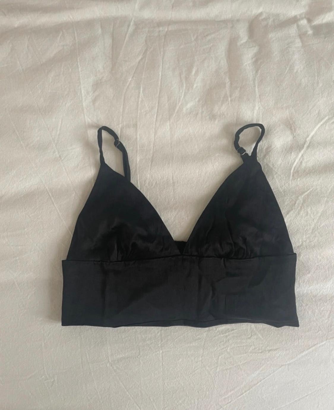 Black bralette with adjustable thin straps and a V-neckline, displayed on a crumpled white sheet. Casual style.