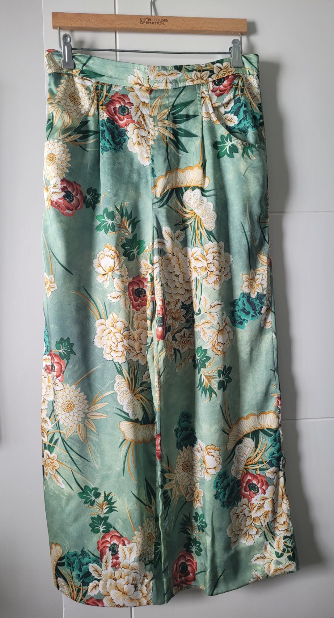 Green fabric trousers with a floral print in red, white, and yellow tones, hanging on a wooden hanger.