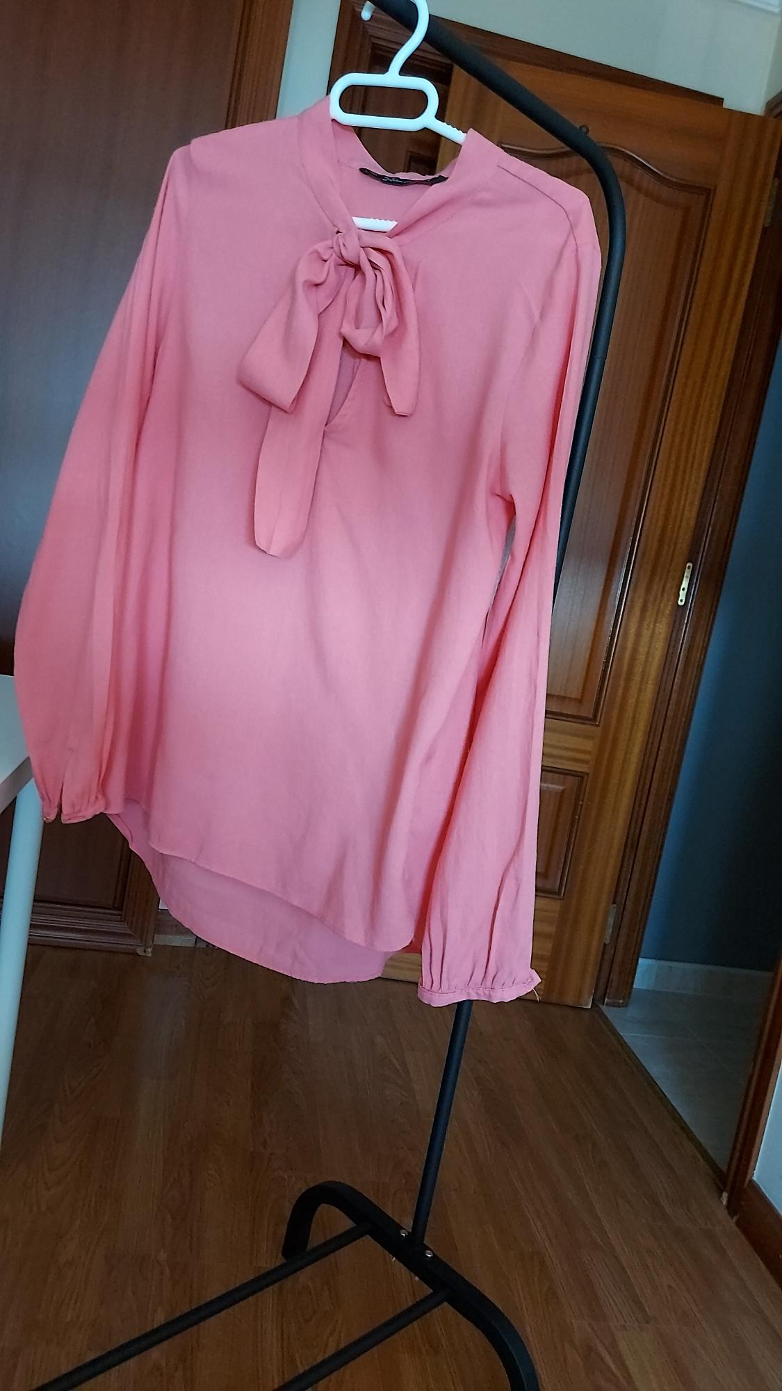 Pink blouse with a bow at the neck, hanging on a black clothes rail, against a background of wooden doors and parquet flooring.