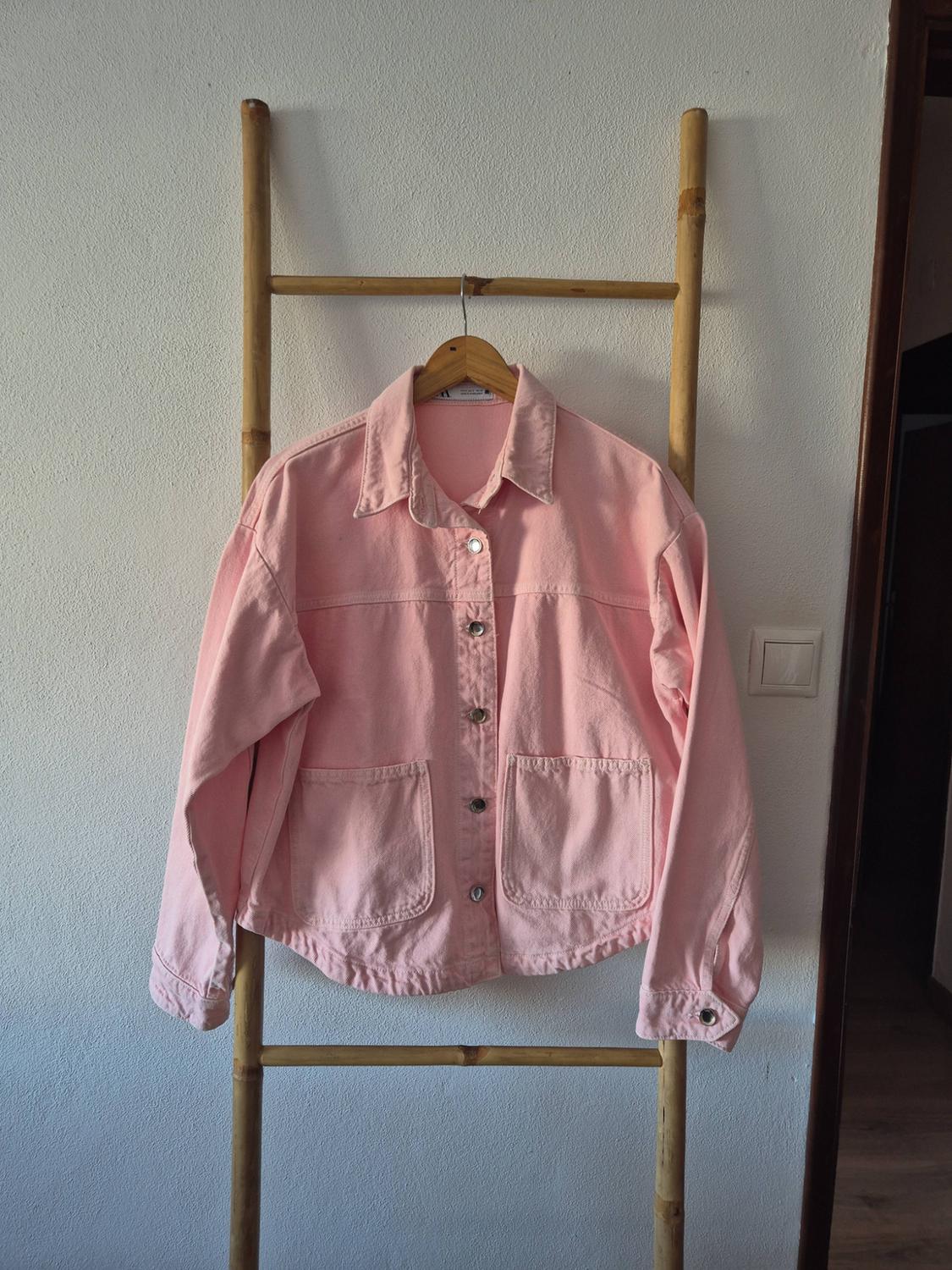 Pink jacket with buttons and pockets, hanging on a hanger on a wooden ladder leaning against a white wall.