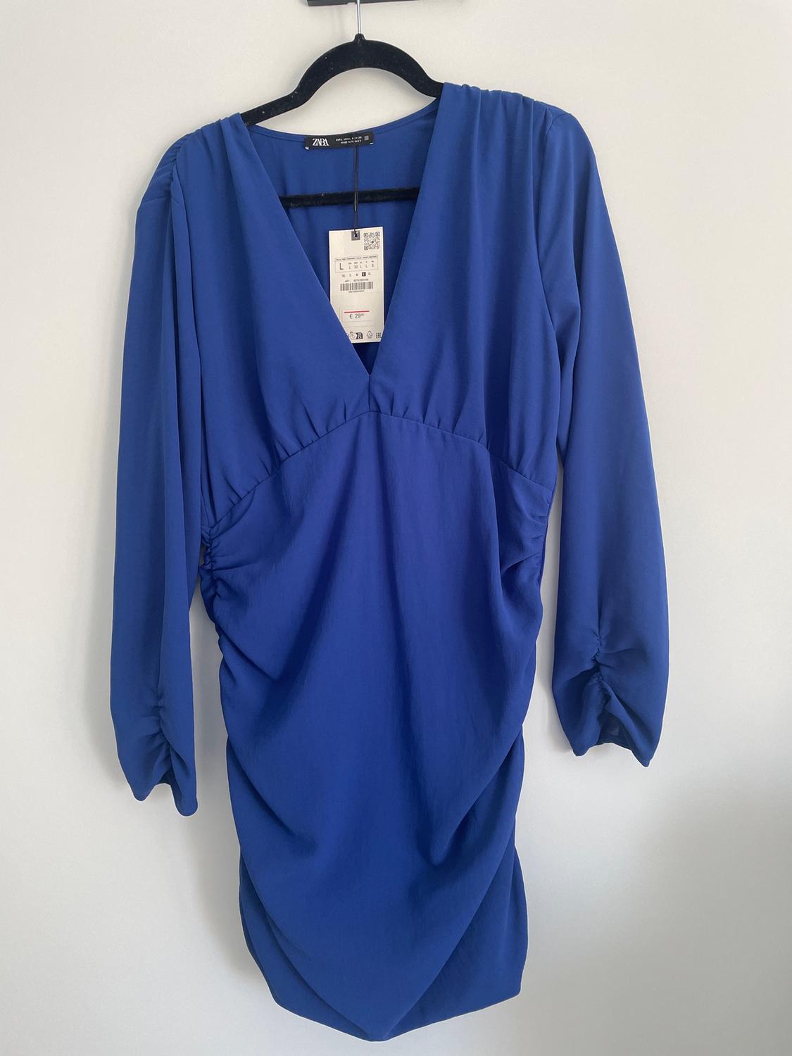 Blue dress with a V-neckline and long sleeves, hanging on a black hanger. Label visible on the collar.