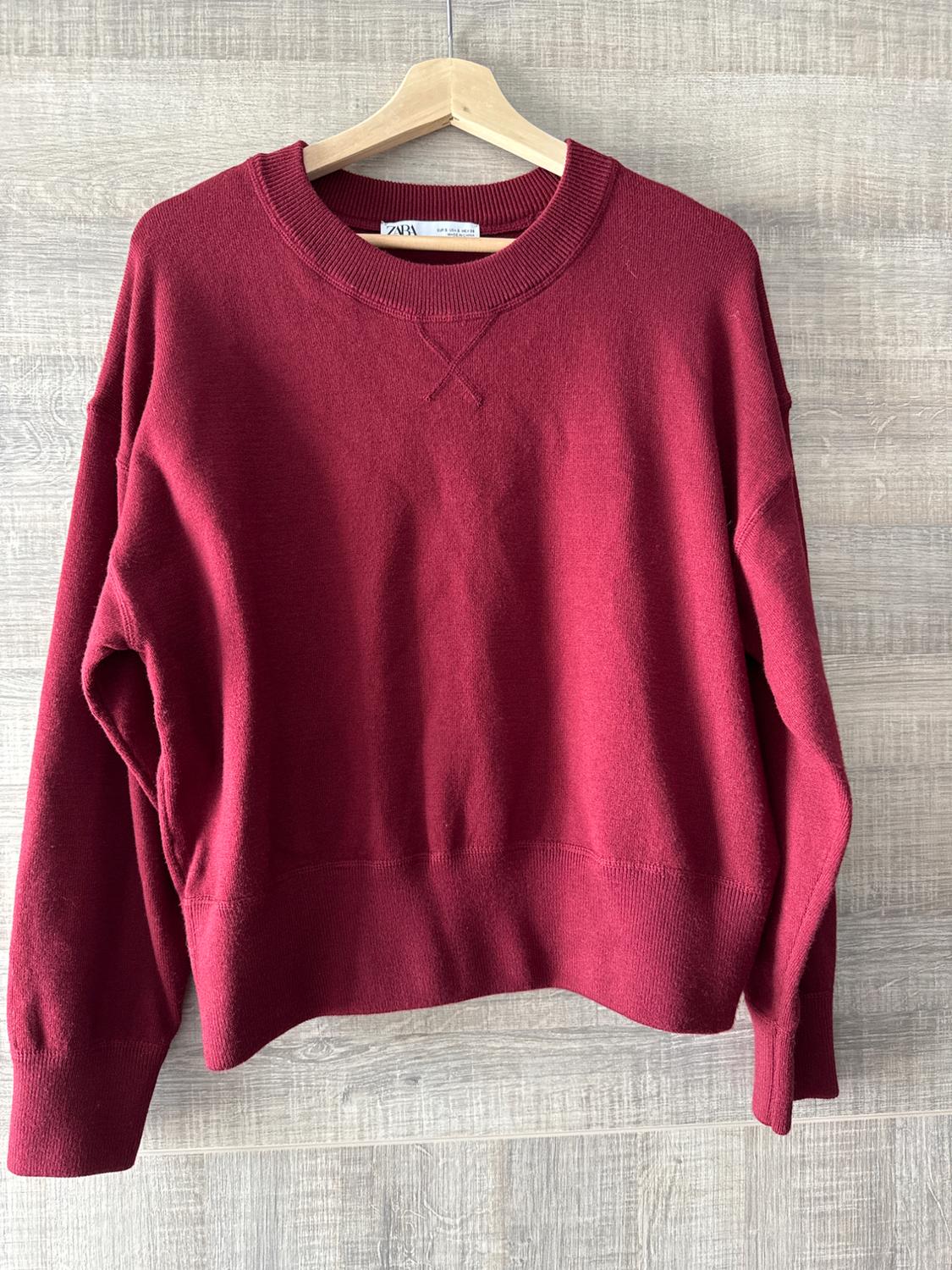 Red long-sleeved jumper hanging on a wooden hanger against a light wood background. Label visible at the neck.