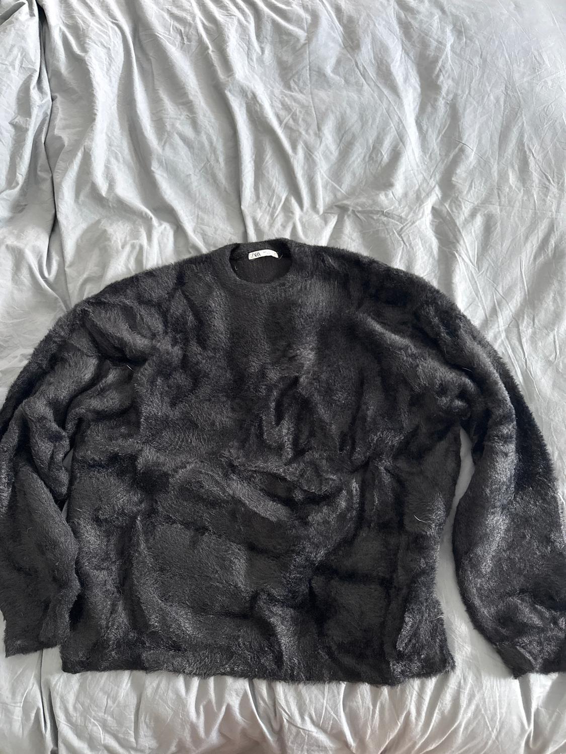 Black fleece jumper on a bed with crumpled grey sheets. Visible label on the jumper's collar.