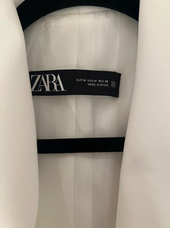 White garment label with text "ZARA, EUR M, USA M, MEX 28, MADE IN SPAIN" on a black background. Diagonal textured fabric.