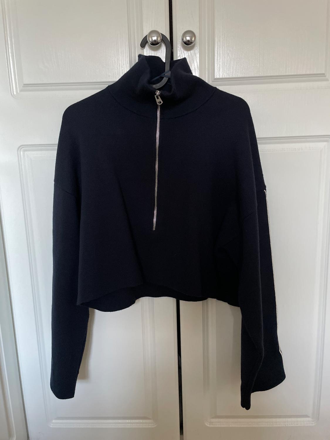 Black cropped long-sleeved sweatshirt with a half-zip front, hanging on a hanger from the silver knob of a white door.