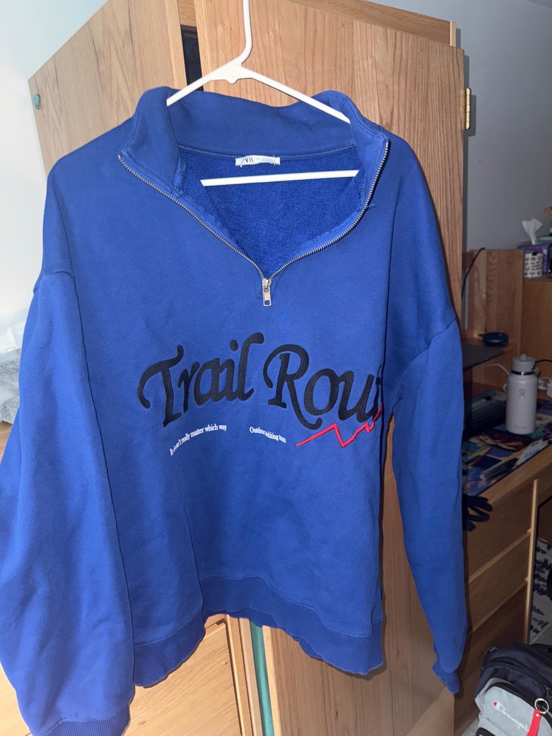 Blue zip-up hoodie with "Trail Route" text, hanging on a white hanger. Background of wooden furniture and various objects.