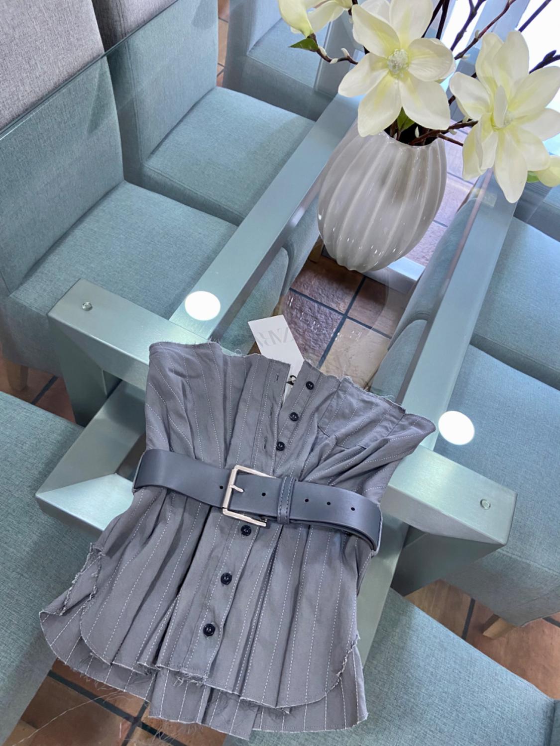 Top view of a grey blouse with a black belt and front buttons on a glass table.