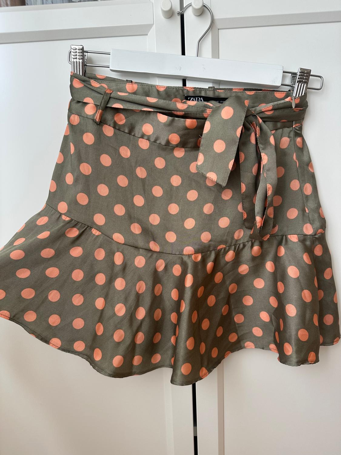 Green mini skirt with pink polka dots and a belt, hanging on a hanger in front of a white wardrobe.
