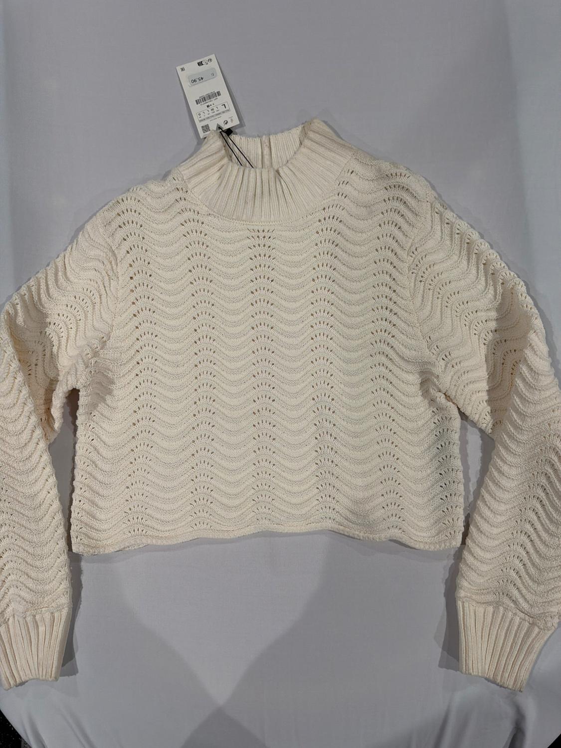 Cream cable-knit jumper with openwork wave pattern, long sleeves and ribbed high neck. Label with text 45.90. Laid flat on light fabric.