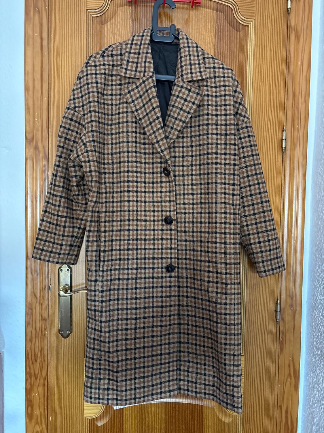 Brown and black checked coat hanging on a wooden door. It has three buttons and a classic design.