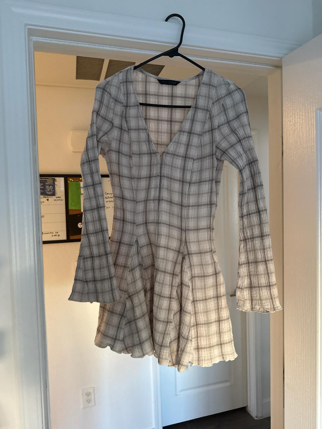 Short light-check dress with a V-neckline and flared sleeves, hanging on a black hanger over the frame of a white door.