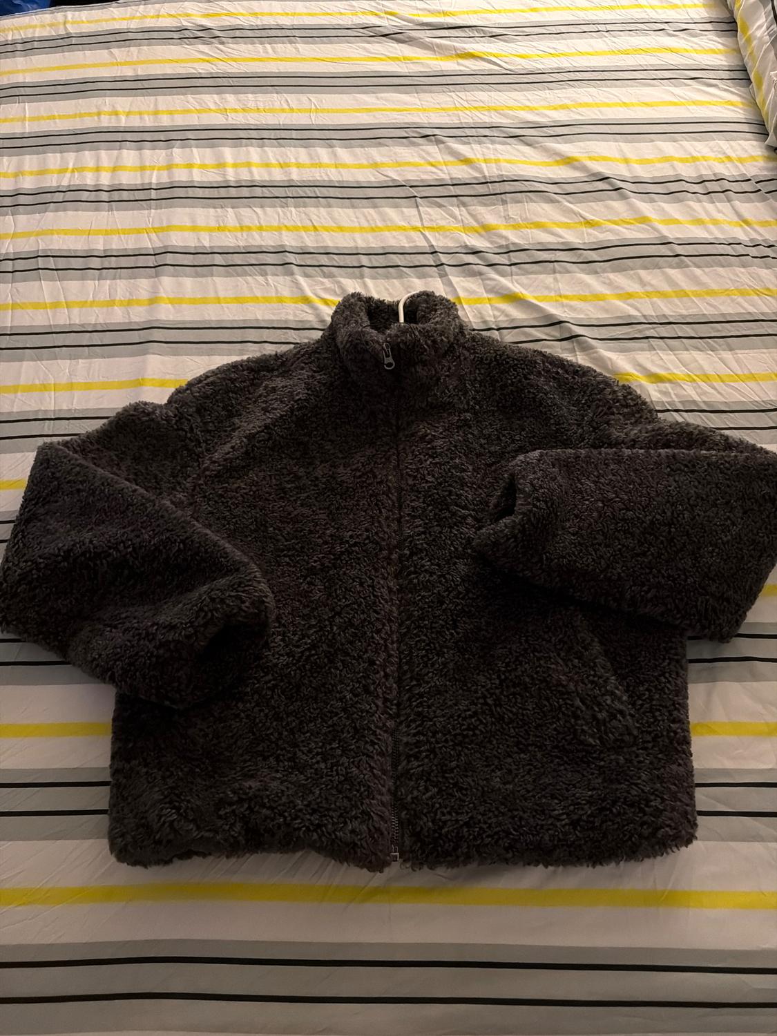 Black fleece jacket with a zip, placed on a bed with grey and yellow striped sheets.