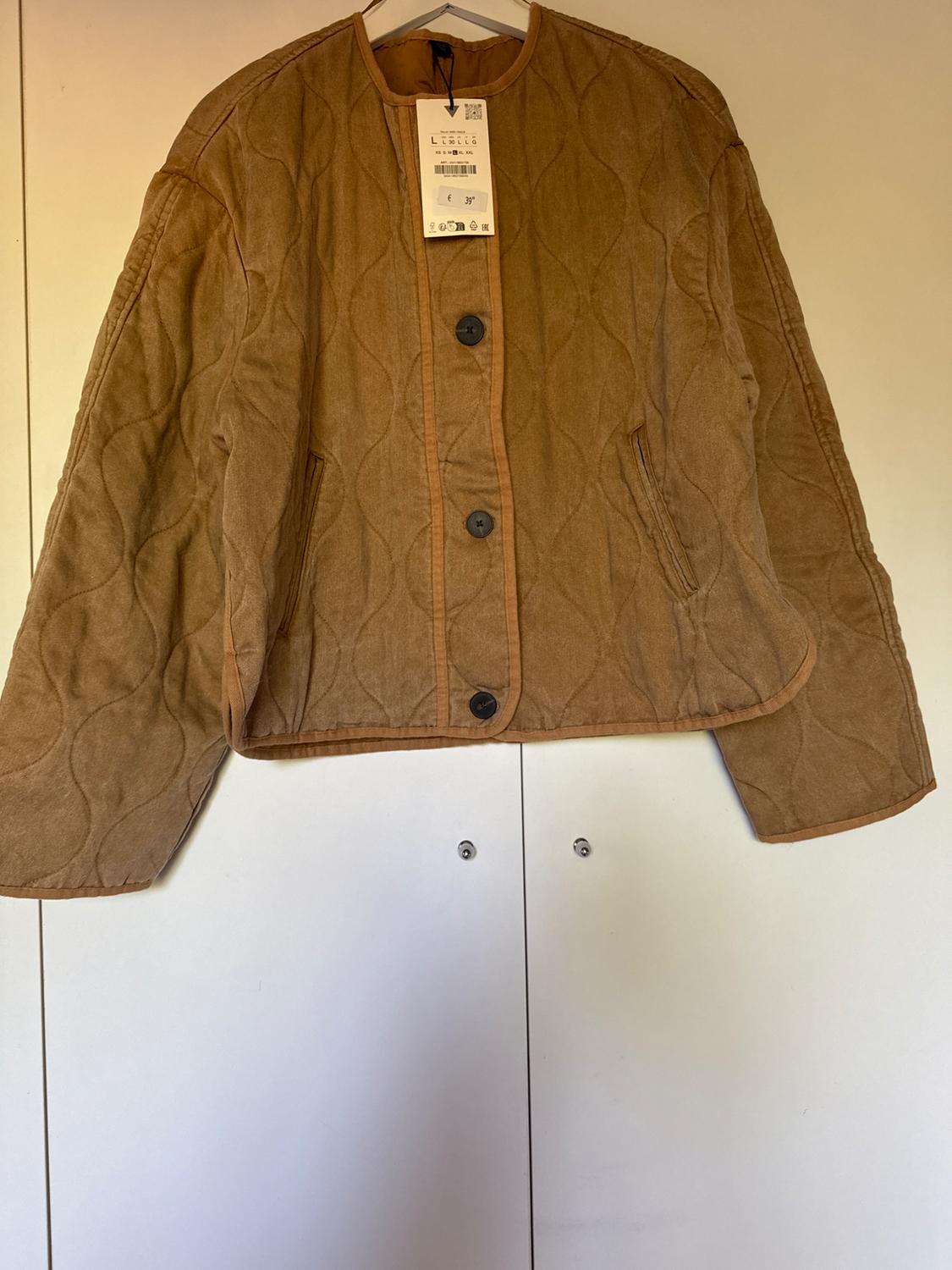 Light brown padded jacket with black buttons and hanging tag, hanging on a hanger against a white background.