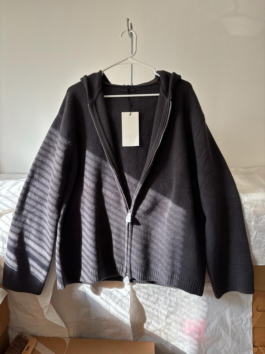 Dark gray knit jacket with hood and zipper on a hanger. Label with ZARA text. Natural lighting with blinds shadows.