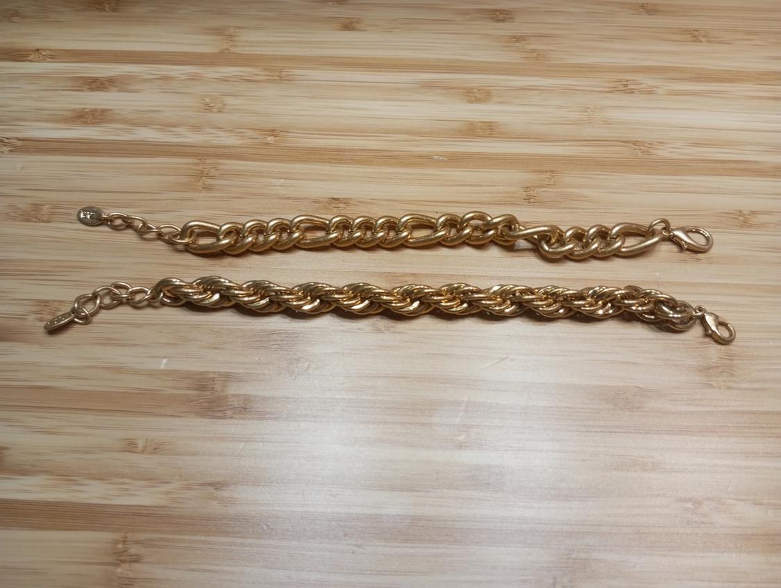 Two gold bracelets on a light wooden surface. One has large links and the other a braided design.