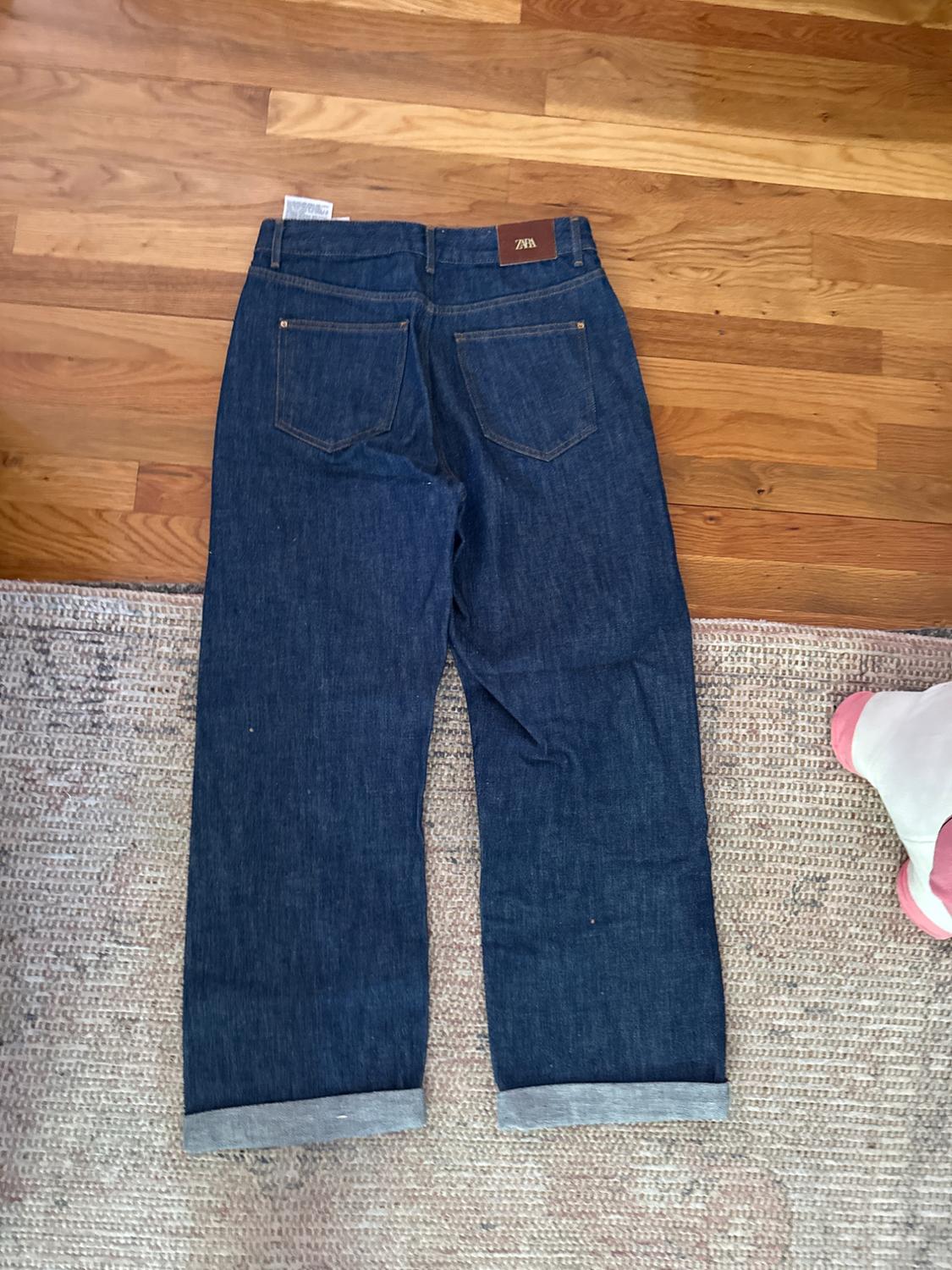 Dark blue denim jeans with rolled cuffs, brown waist tag, on a wooden floor and light carpet.