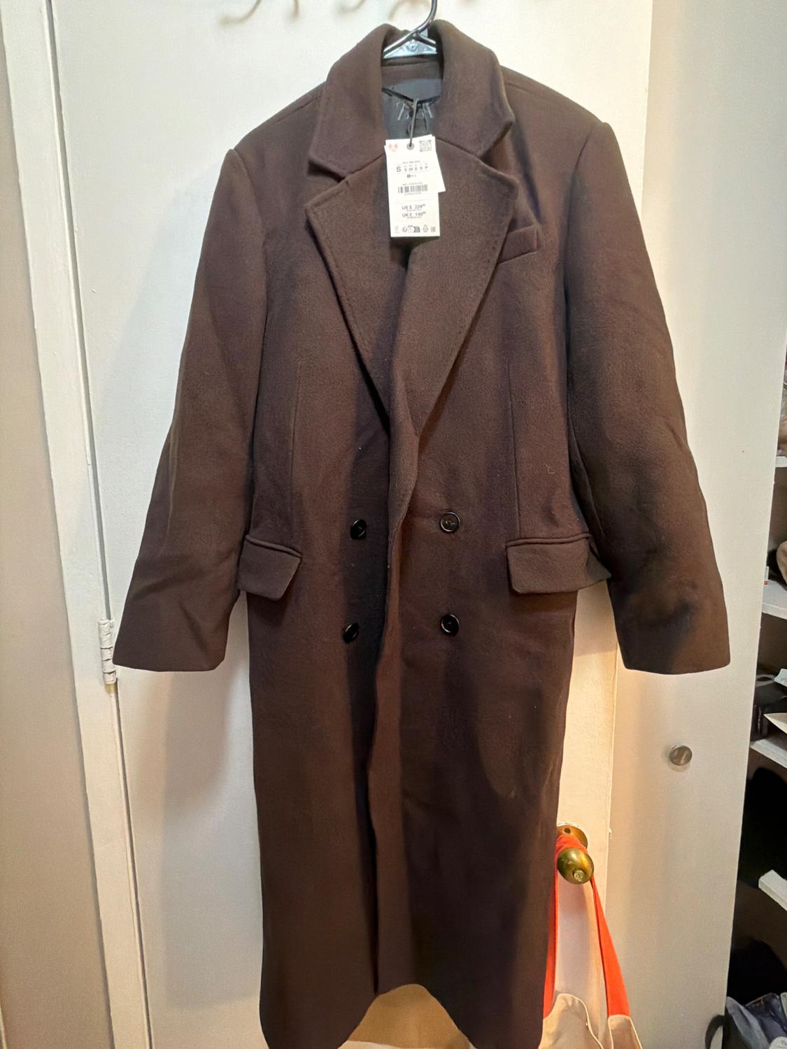 Long brown, belted coat hanging on a white door. It features wide lapels, black buttons, flap pockets, and a tag.