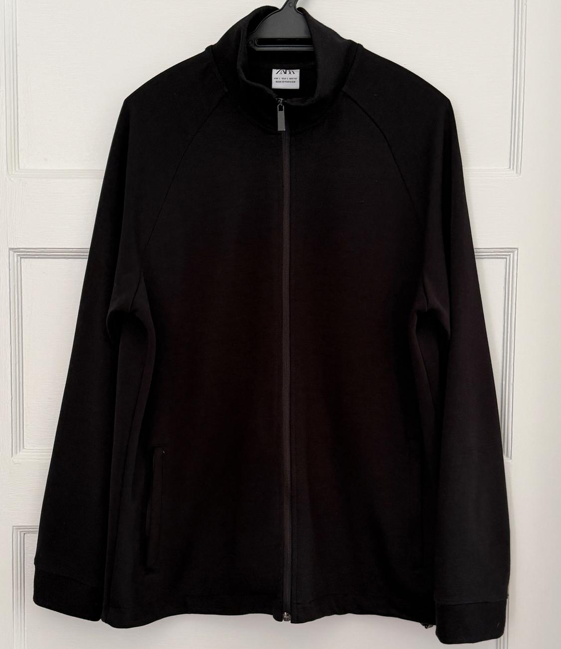 Black long-sleeved jacket with a front zipper, hanging on a hanger over a white paneled door. Label with ZARA text.