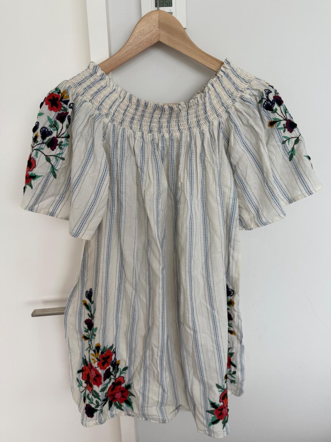 Blue and white striped blouse with floral embroidery on the sleeves and hem, hanging on a wooden hanger over a white door.
