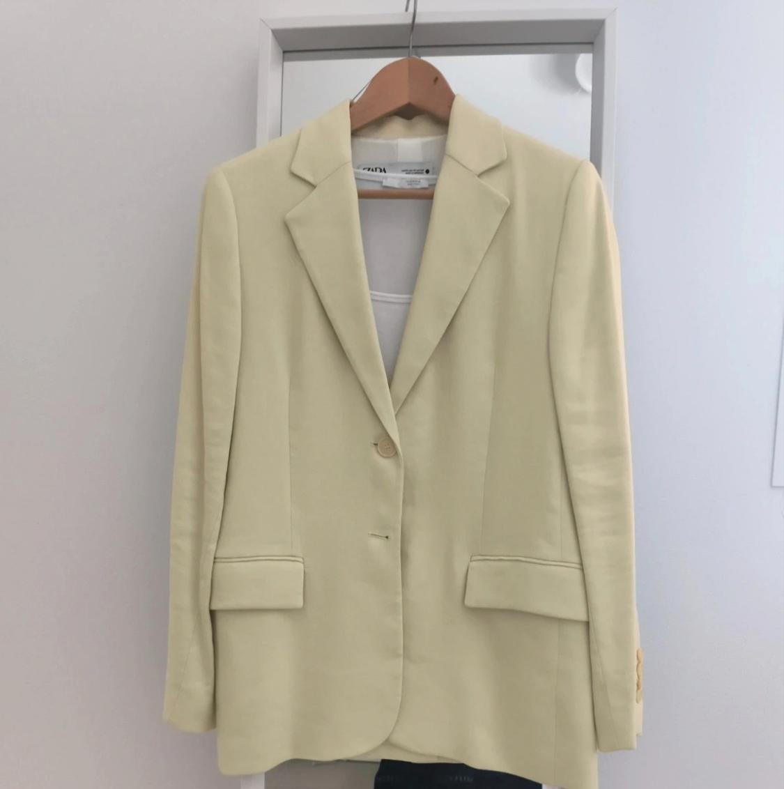 Light yellow button-up blazer with ZARA label, hanging on a wooden hanger in front of a mirror on a white wall. Minimalist setting.