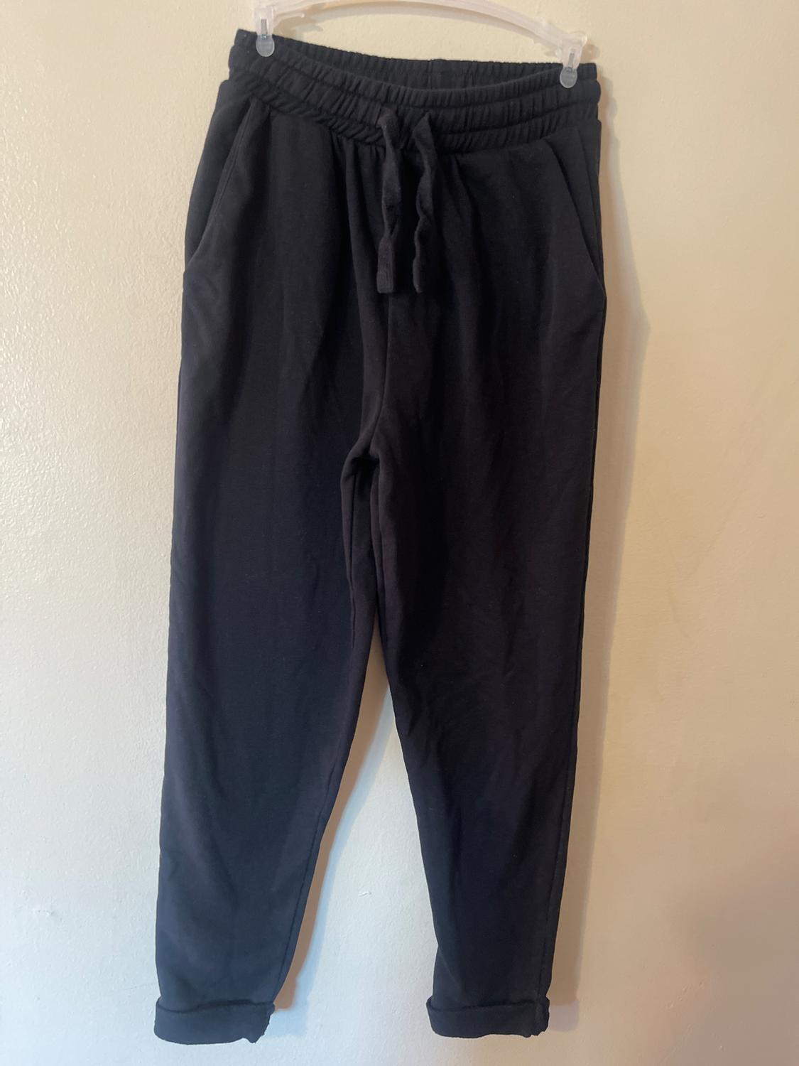 Black pants hanging on a clear hanger, with an elastic waistband and adjustable drawstring, against a light beige background.