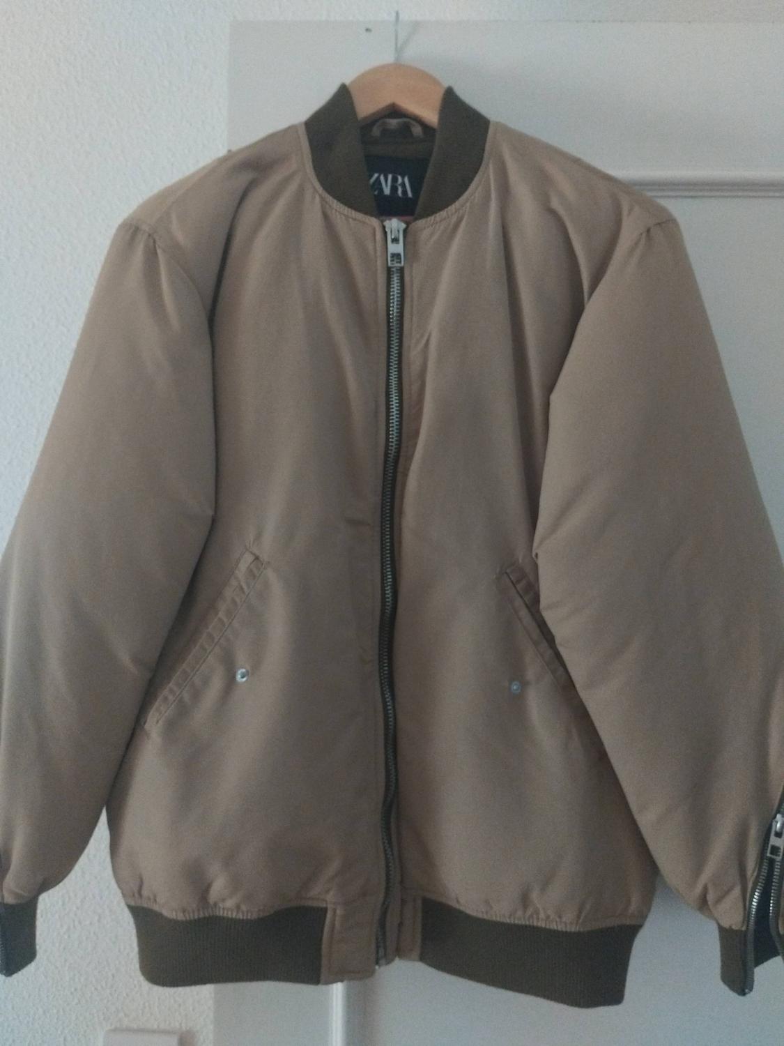Beige bomber jacket on a hanger over a white door. Front zip, dark collar and cuffs. Inner label with the text ZARA.