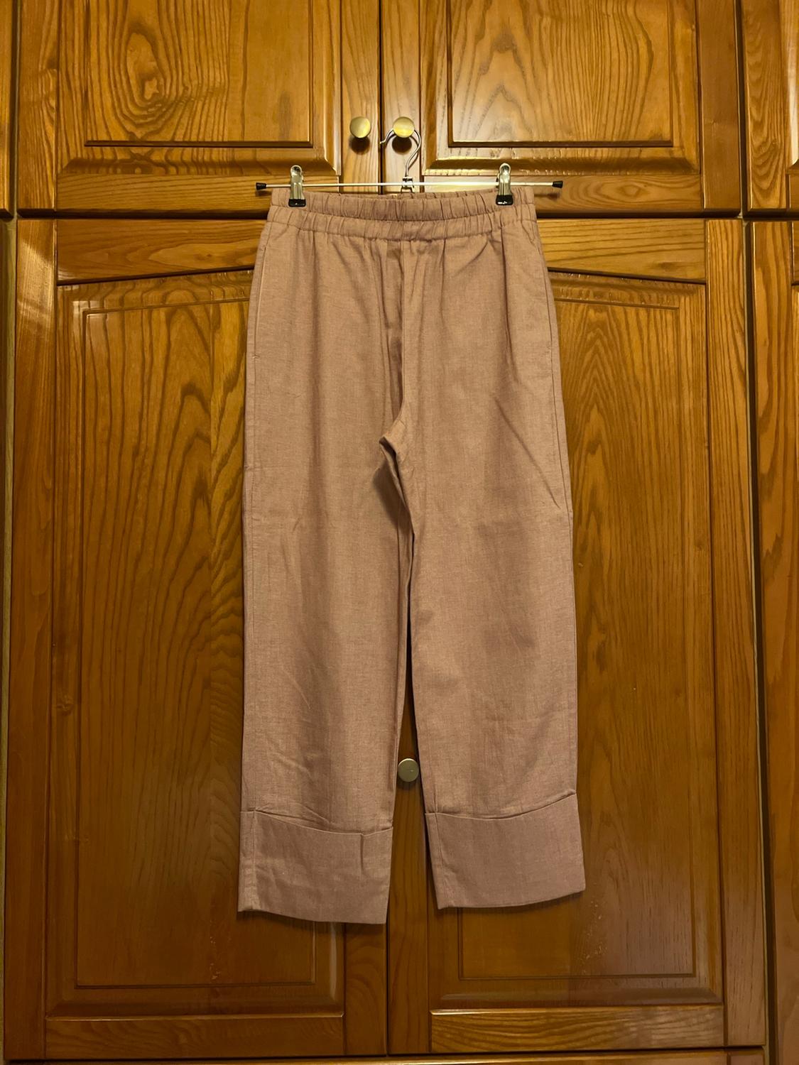 Light pink trousers with an elasticated waist and turn-up hems, hanging on a coat hanger in front of some wooden wardrobe doors.