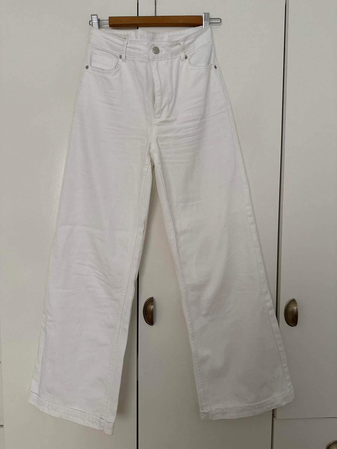 White, wide-leg trousers hanging on a wooden hanger in front of a white wardrobe with gold handles.
