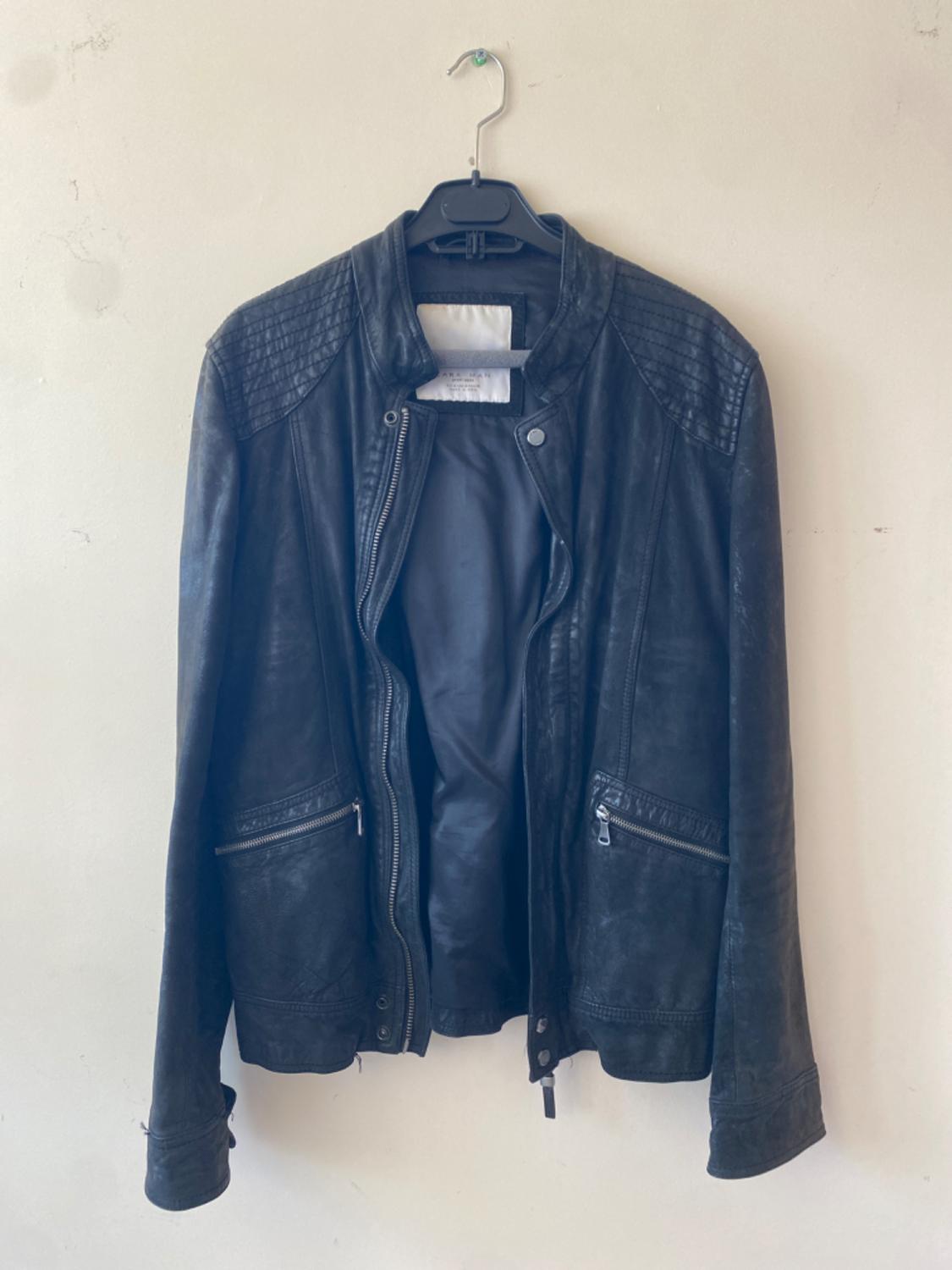 Black leather jacket hanging on a hanger against a white wall. It has zips on the pockets and a quilted design on the shoulders.