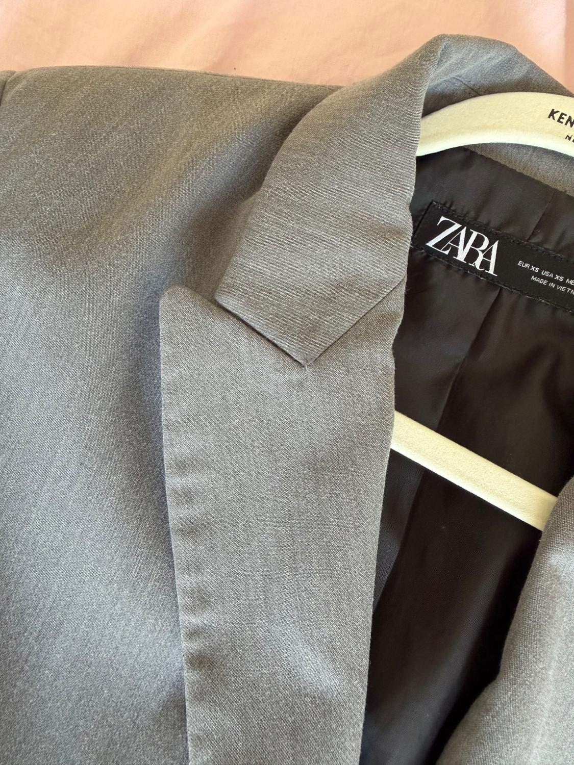 Close-up of a grey blazer on a white hanger against a pink background. Showing lapel, black lining and a label with the text ZARA EUR XS USA XS.
